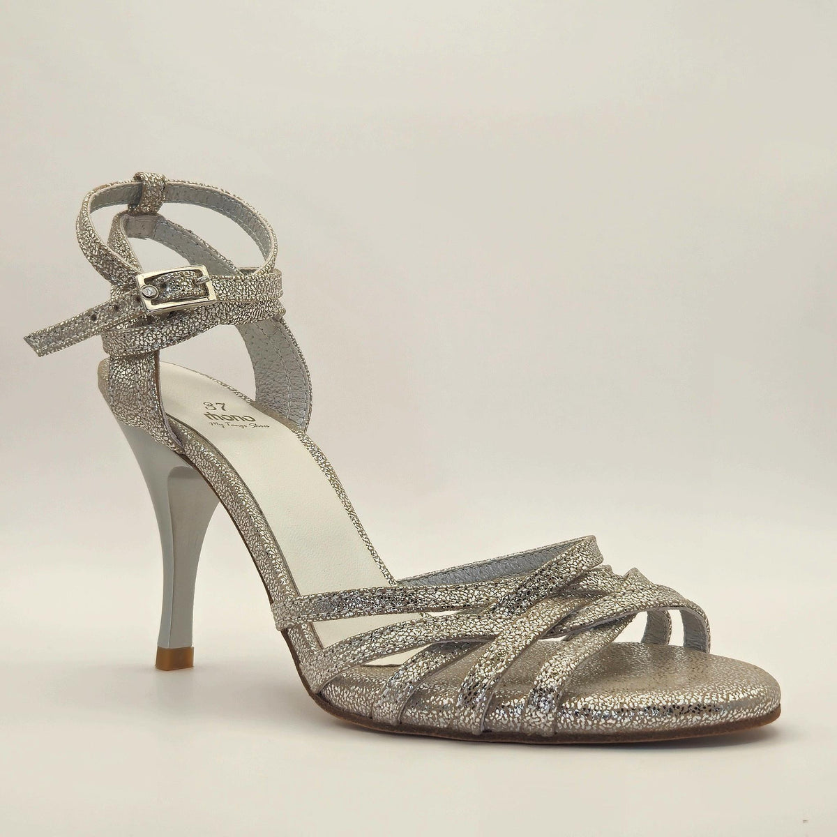 Three-quarter front view of Mono Lunar Glass tango sandal with the long strap in a single ankle wrap and square silver buckle, showing the multi-strand forefoot in iridescent silver crinkle leather, white leather insole with Mono branding, and pearl-white stiletto heel.