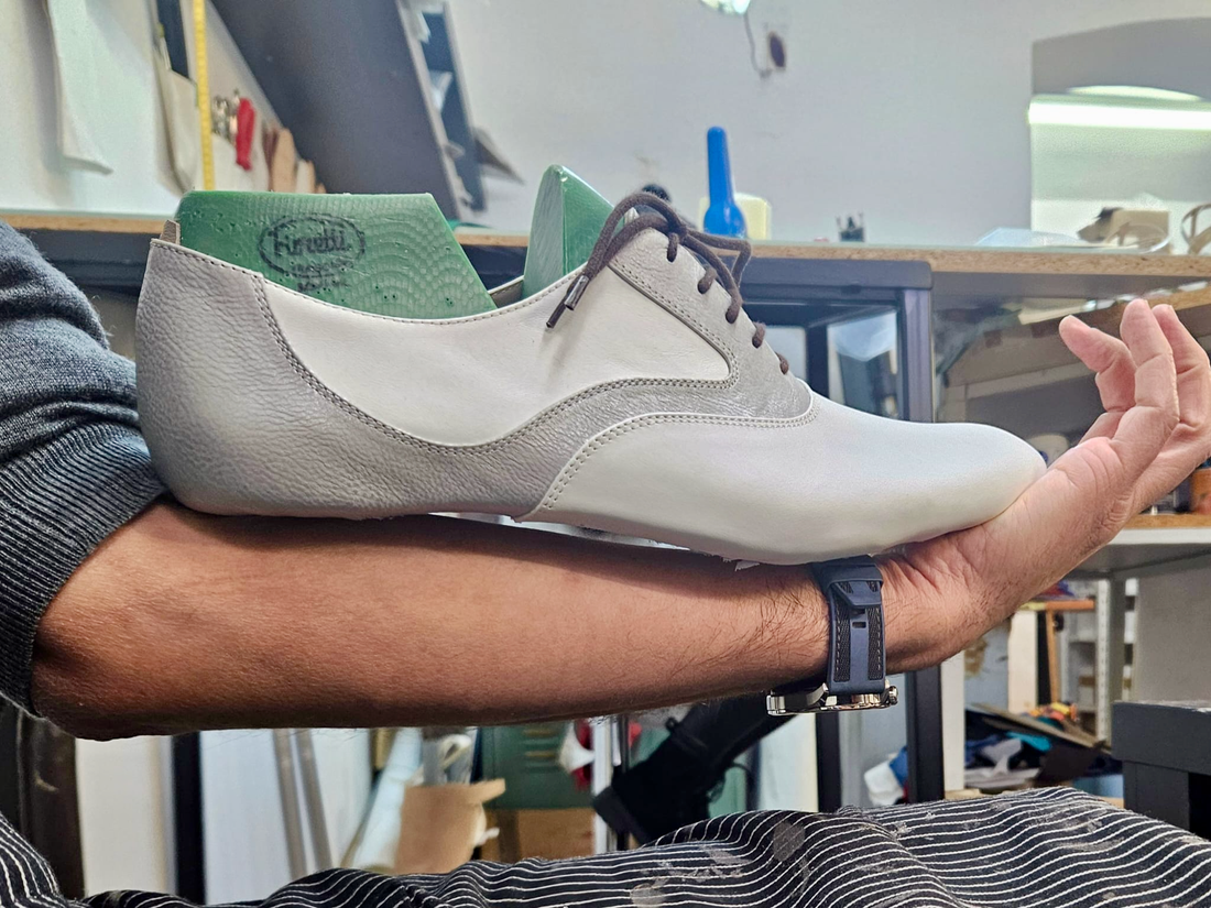 A finished men's Mono tango shoe in white and gray leather held up by a shoemaker's arm in the Timișoara atelier, with shoe lasts and workshop tools visible in the background
