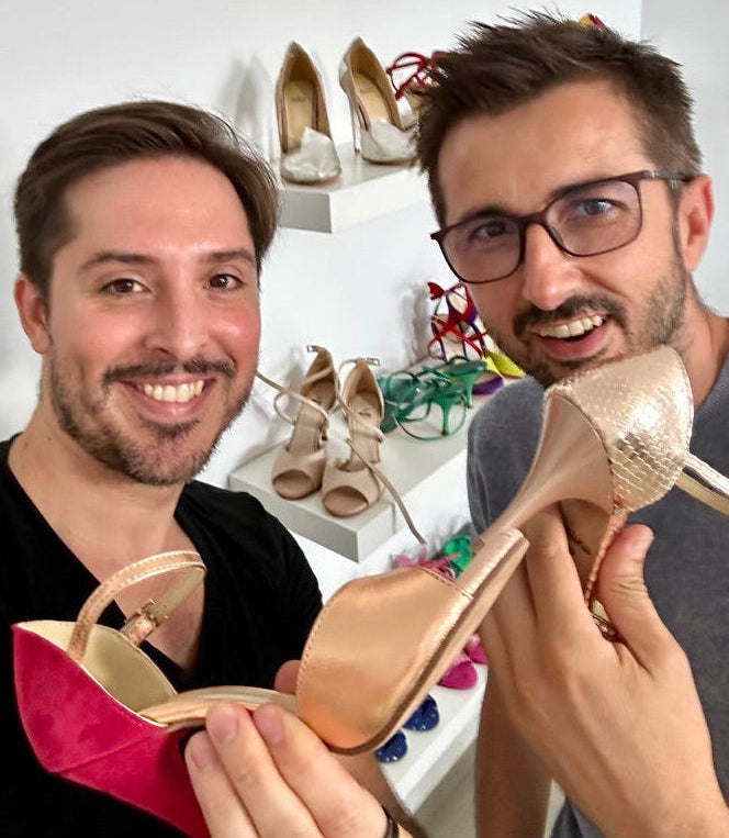 Two men smiling and holding high-heeled shoes in a room with shelves of other shoes in the background. One shoe is a metallic champagne beige with a textured heel, and the other is a bright pink with a beige upper.