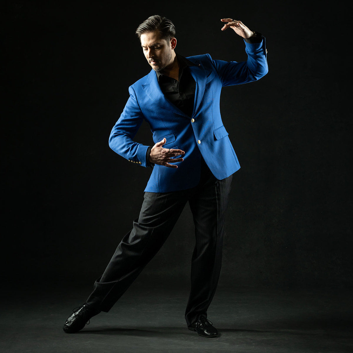 A man in a bright blue blazer and black trousers poses dynamically against a solid black studio background.