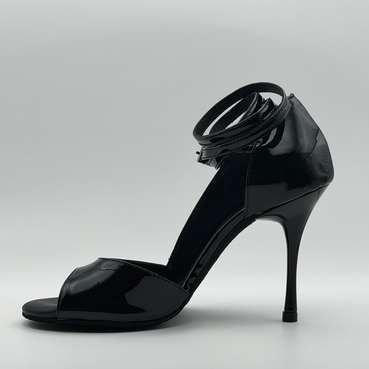 Inner side view of the Akvile Noir black patent leather tango heel showing the open side, closed heel counter, and wrapped ankle strap