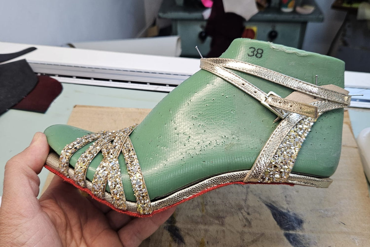 A handmade Mono tango shoe mid-construction on a wooden last in the Timișoara atelier, showing the green satin upper, crystal-embellished ankle straps, and buckle closure before the final finishing.