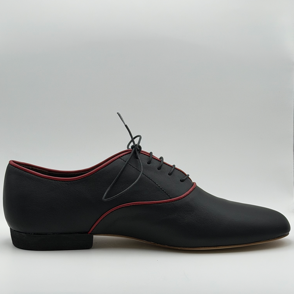 Side profile of the Mono El Hilo Rojo men's tango oxford in black leather, showing the fine red piping tracing the topline and panel curves