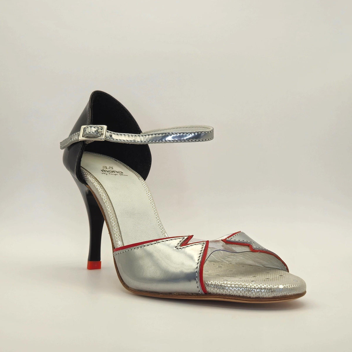 Side profile of Mono Electra Glam women's tango shoe with closed heel cage facing left, showing the metallic silver and red-trimmed vamp, glossy black patent heel counter, and slender stiletto heel.