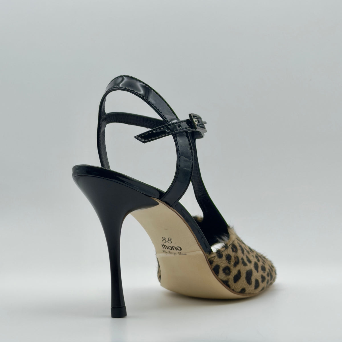 High rear three-quarter view of Mono La Fiera tango sandal showing the back of the T-bar, black patent ankle strap with silver buckle, the full curved stiletto heel, and the natural leather outsole stamped with the Mono logo and size.