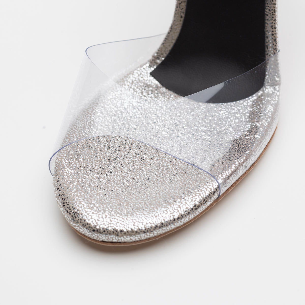 Close-up detail of Mono La Plata closed-heel tango shoe toe showing the clear PVC vamp, silver glitter toe bed texture, and black fine-grid textured closed heel counter in the background.