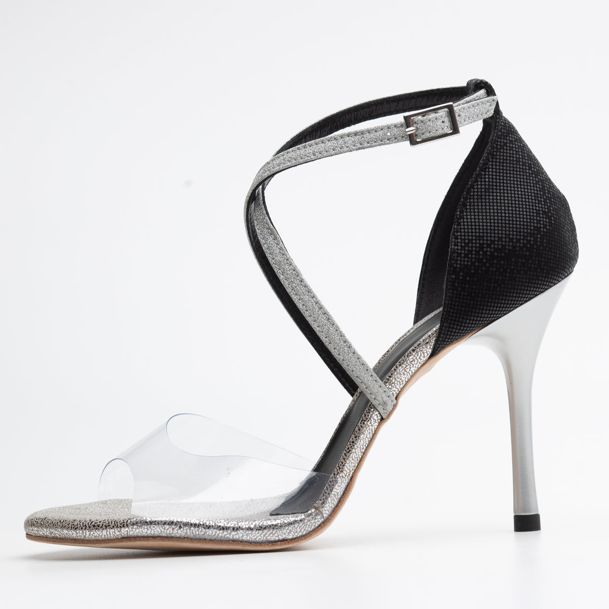 Side profile of Mono La Plata women's closed-heel tango shoe facing left, showing the clear PVC vamp over a silver glitter toe bed, silver glitter crisscross ankle straps, black fine-grid textured closed heel counter, buckle closure, and a slender silver stiletto heel.