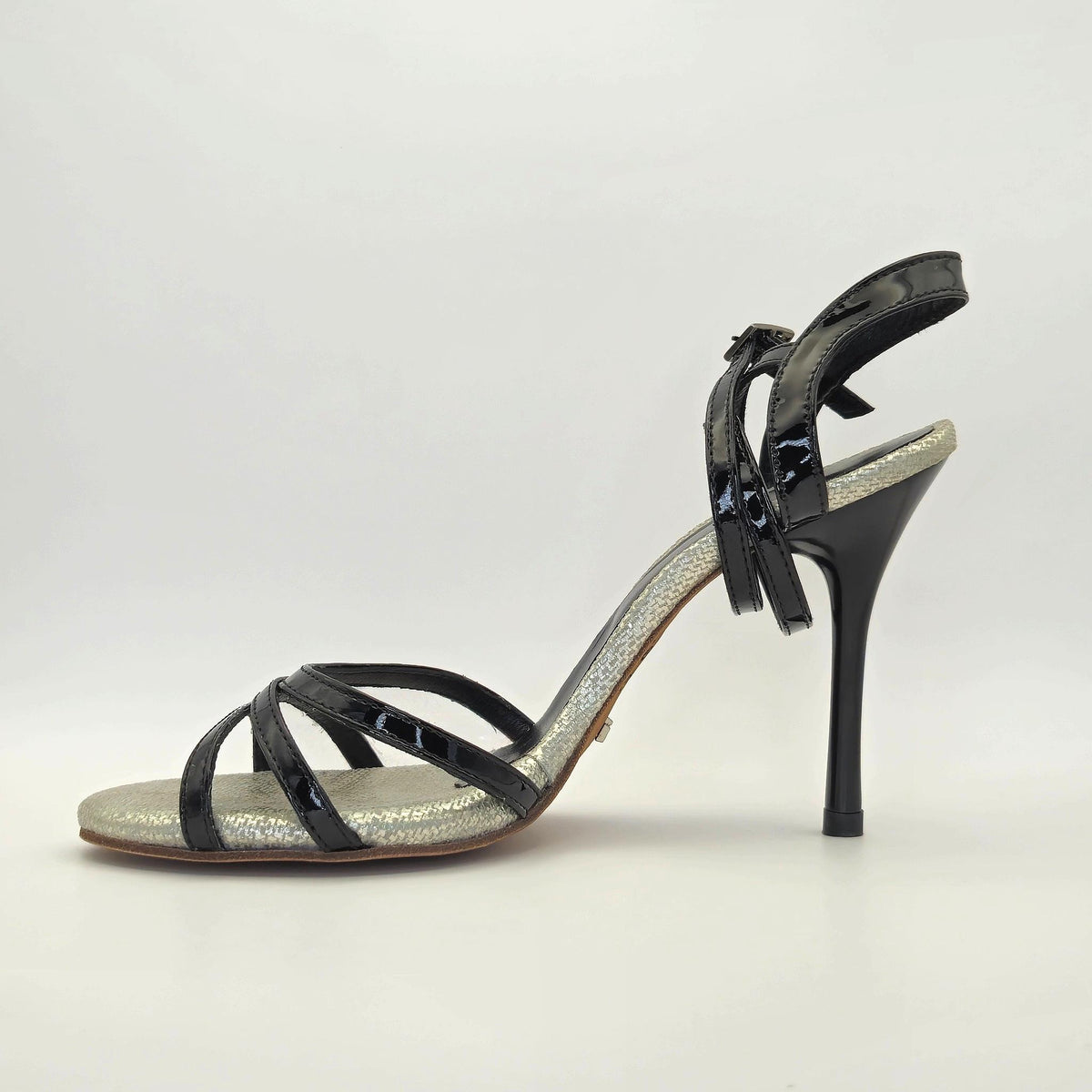 Left side profile of Mono Midnight Glitz tango sandal with the slingback ankle strap loosely looped at the heel, showing the layered high-gloss black patent cross forefoot straps, silver glitter footbed edge, and the clean arc of the black stiletto heel.