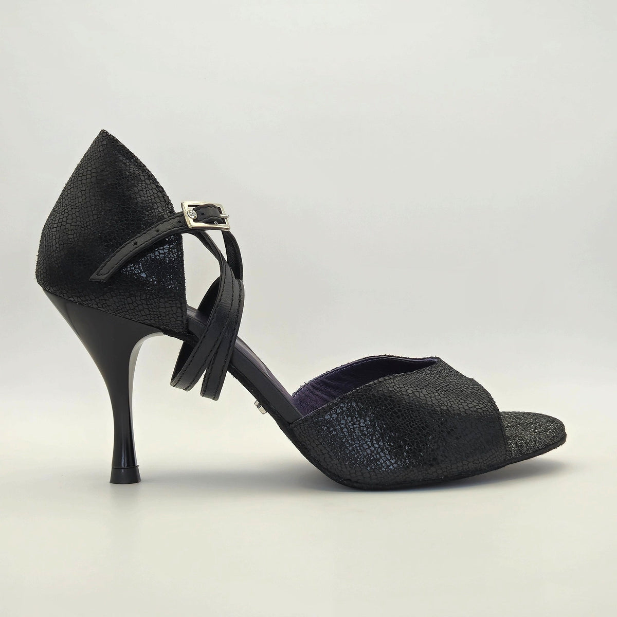 Side profile of Mono Midnight Mystique women's closed-heel tango shoe facing right, showing the black shimmer textured leather closed heel counter, crossover ankle strap with bow, and glimpse of violet insole.
