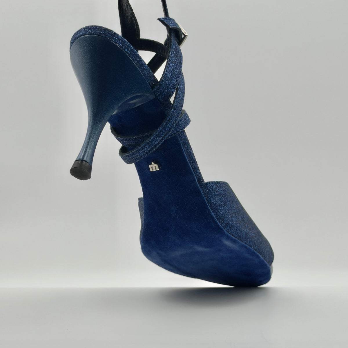 Suspended aerial view of the Mono Sapphire tango sandal sole, revealing the blue suede insole, Mono logo hardware on the heel, and glitter leather upper with crossover ankle straps.