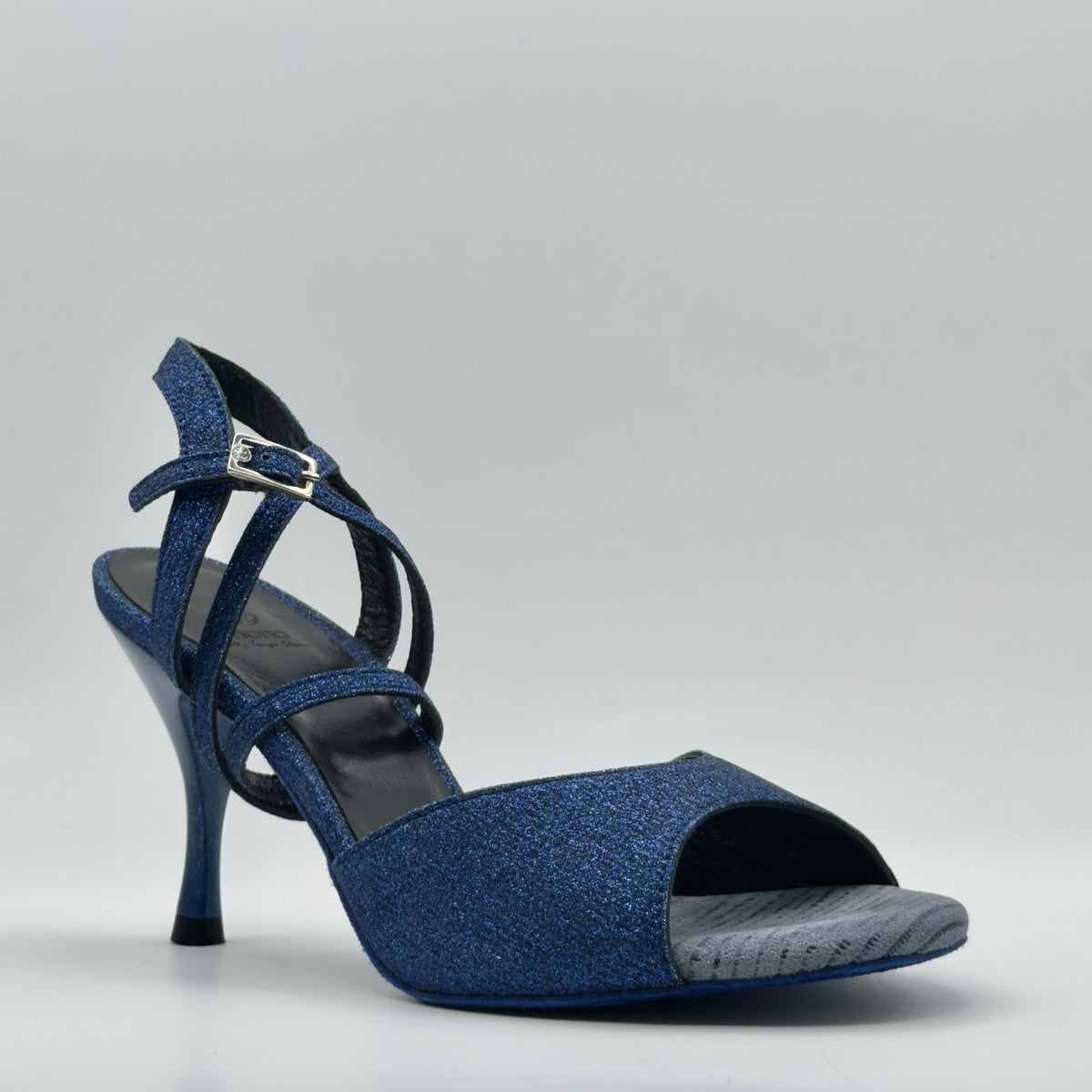 Three-quarter front view of the Mono Sapphire tango heel, showcasing deep blue glitter leather, a wide toe strap, crossover ankle straps, and a sleek stiletto heel against a light gray background.